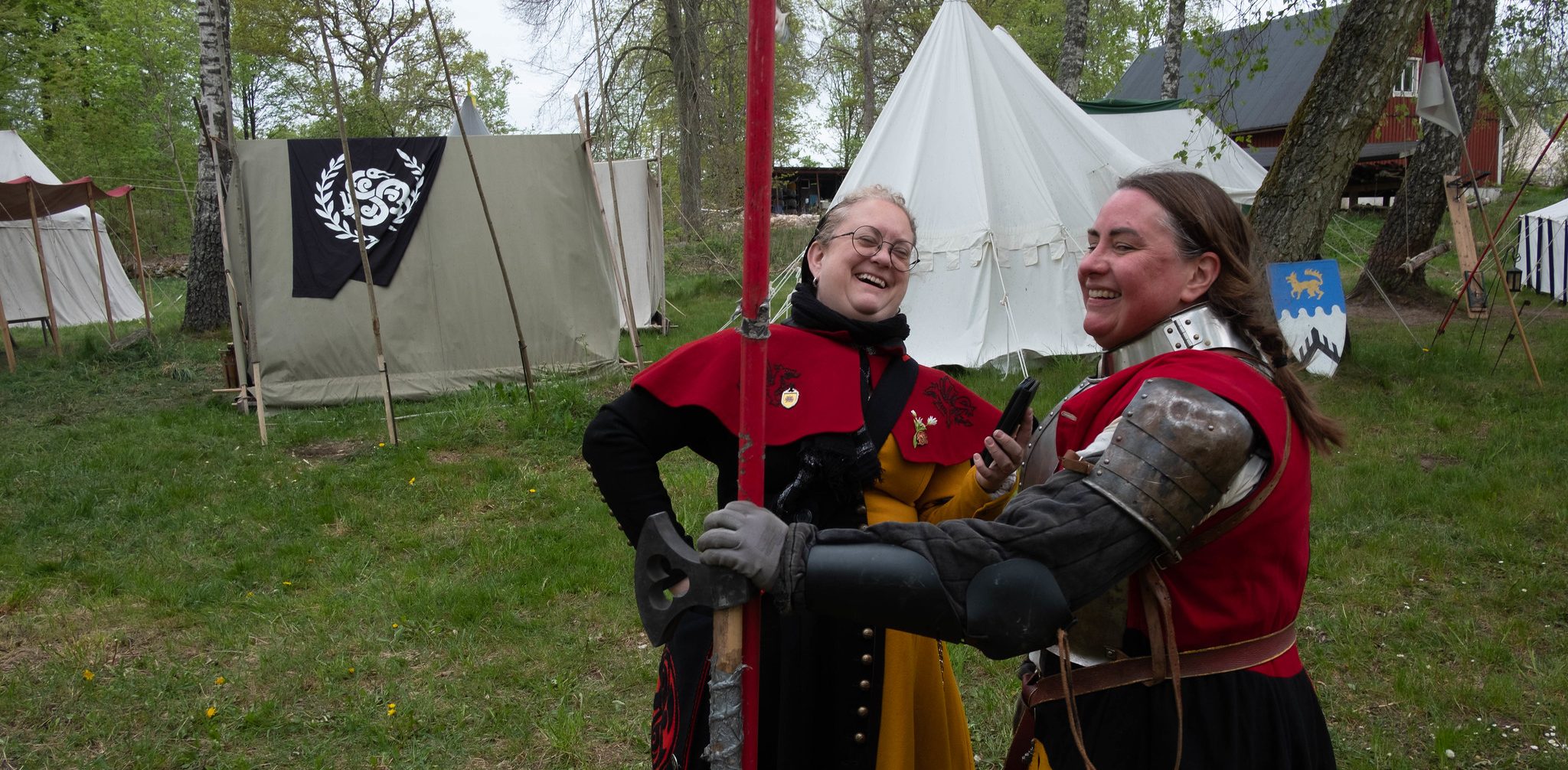 Two women in black and red medival clothes laughing. One is wearing armour and look sweaty from fighting.